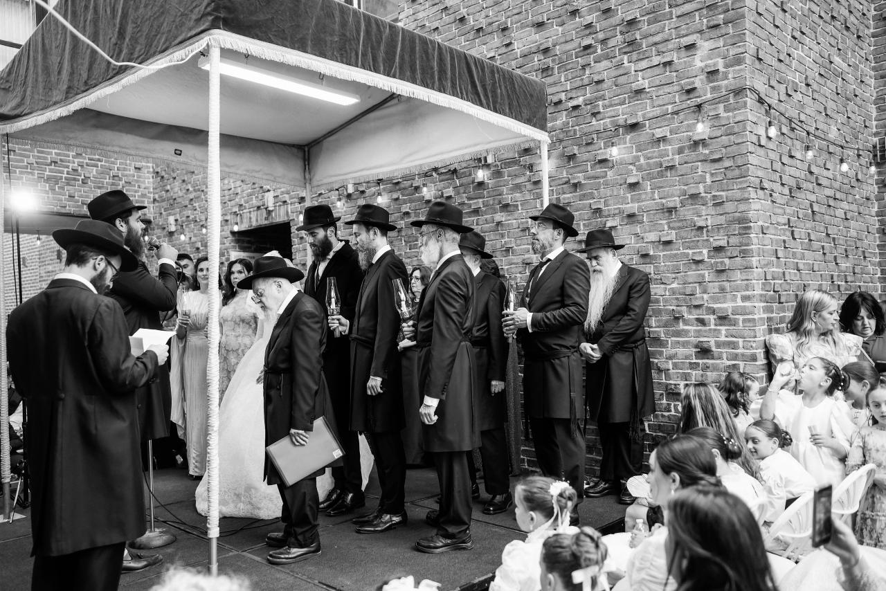 Black-and-white view of the wedding party beneath an outdoor chuppah, with guests seated beside a brick facade. From this Oholei Torah Ballroom wedding. Wedding photography.