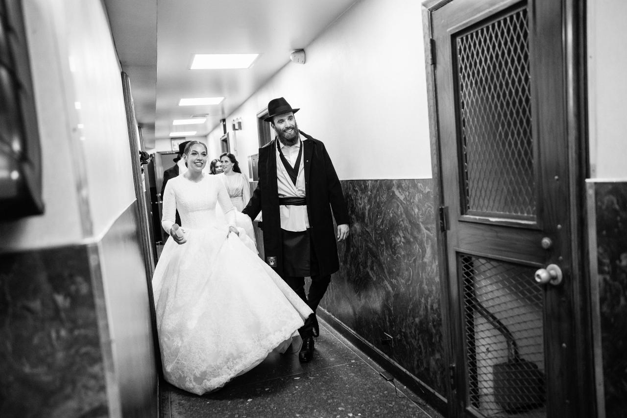 Black-and-white candid of the smiling bride and groom walking hand in hand through a hallway after the ceremony. From this Oholei Torah Ballroom wedding. Wedding photography.