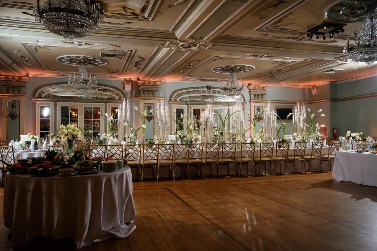 Oholei Torah Ballroom set for a wedding reception, with chandeliers, white flowers, tall glass candles and warm pink uplighting. Wedding photography.