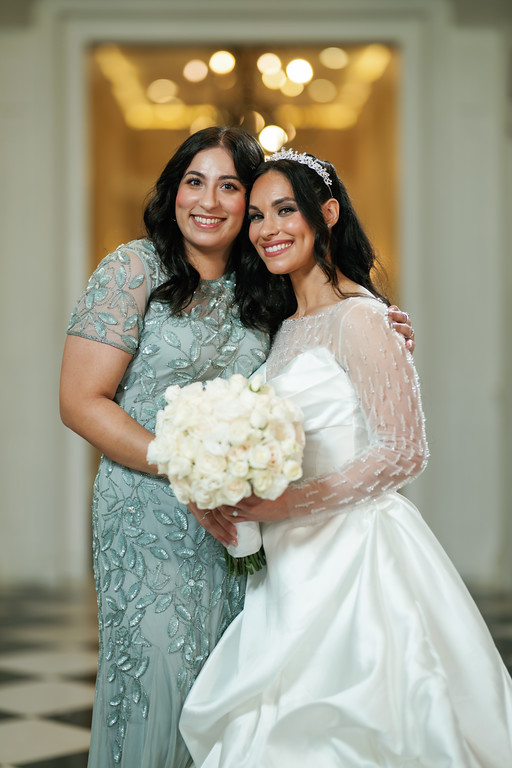 Two women in formal dresses smile while hugging closely at a wedding, with soft golden lights in the background and a bouquet held in the foreground.
