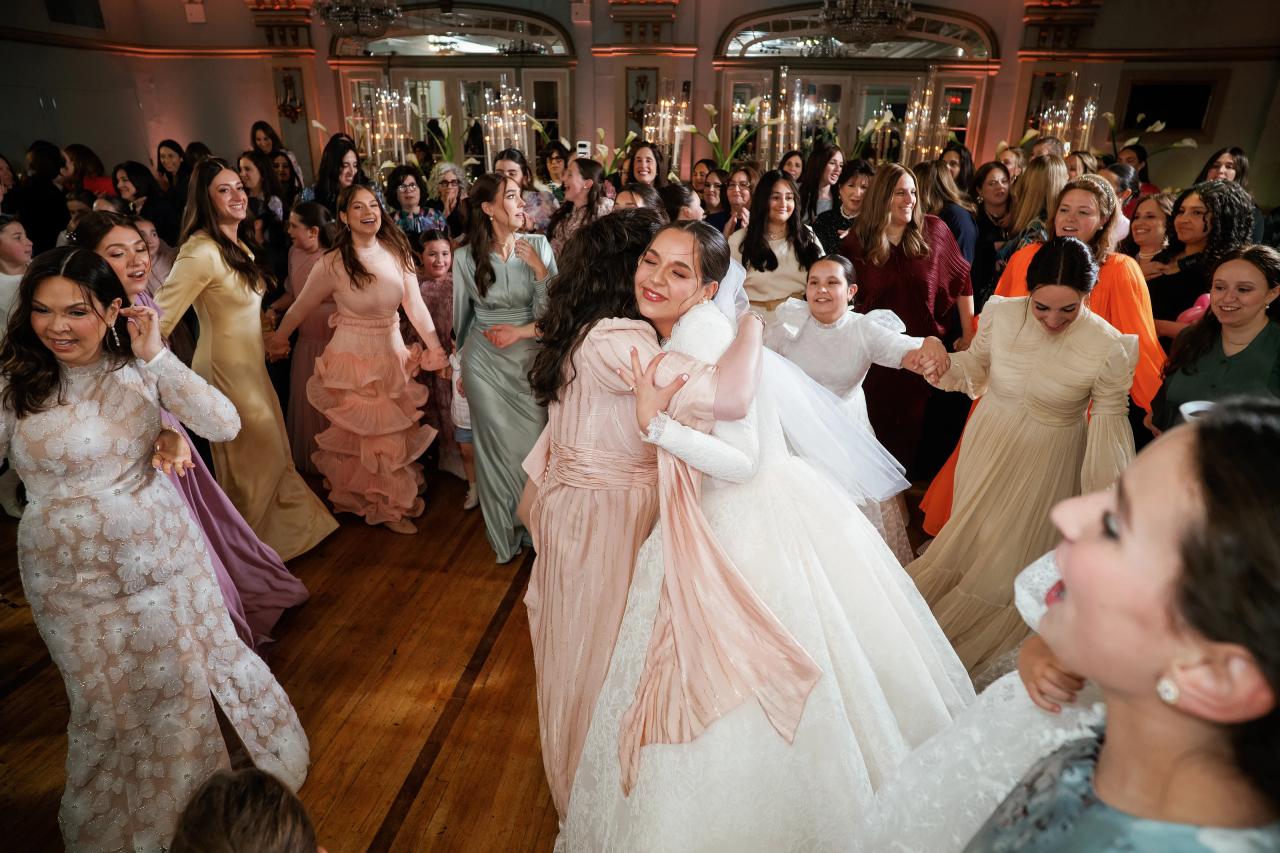 The bride embraces a woman in a pale pink dress as guests sing and dance around them. From this Oholei Torah Ballroom wedding. Wedding photography.