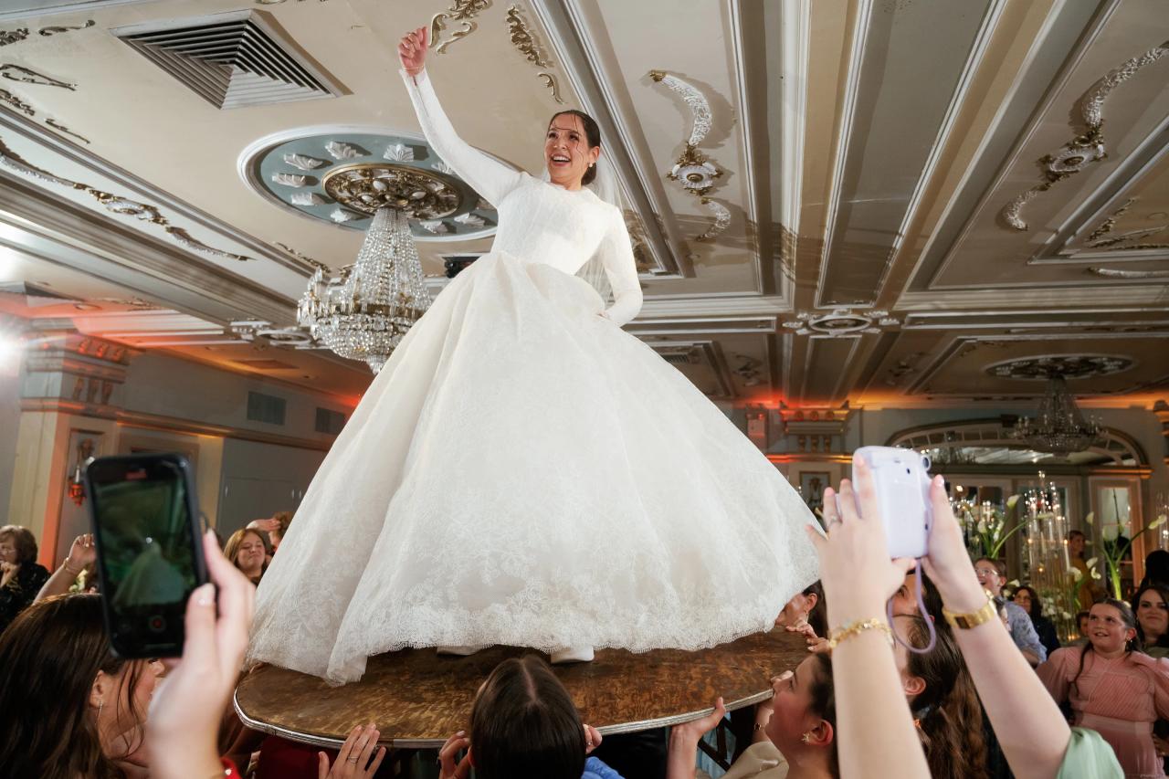 The bride smiles and lifts one arm while standing on a round platform held above celebrating guests beneath an ornate ballroom ceiling. From this Oholei Torah Ballroom wedding. Wedding photography.