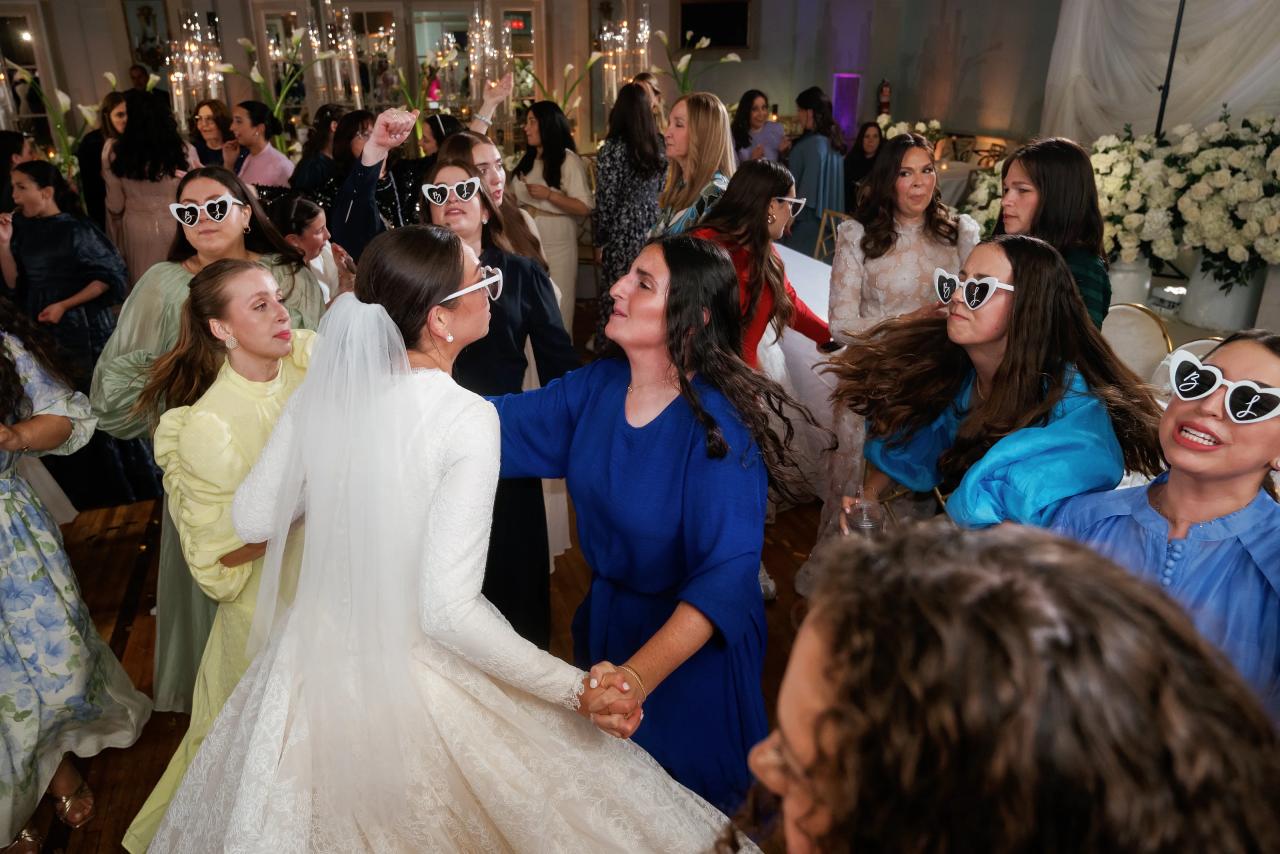 The bride holds hands with a woman in a blue dress while guests dance around them wearing white heart-shaped sunglasses. From this Oholei Torah Ballroom wedding. Wedding photography.