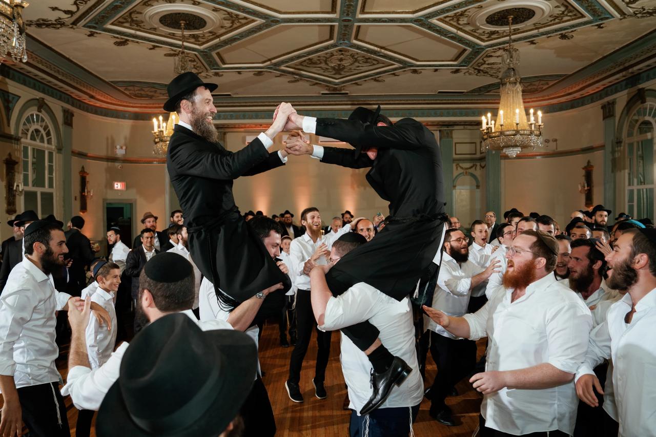The groom and another man hold hands while guests lift them above the wedding dance floor. From this Oholei Torah Ballroom wedding. Wedding photography.