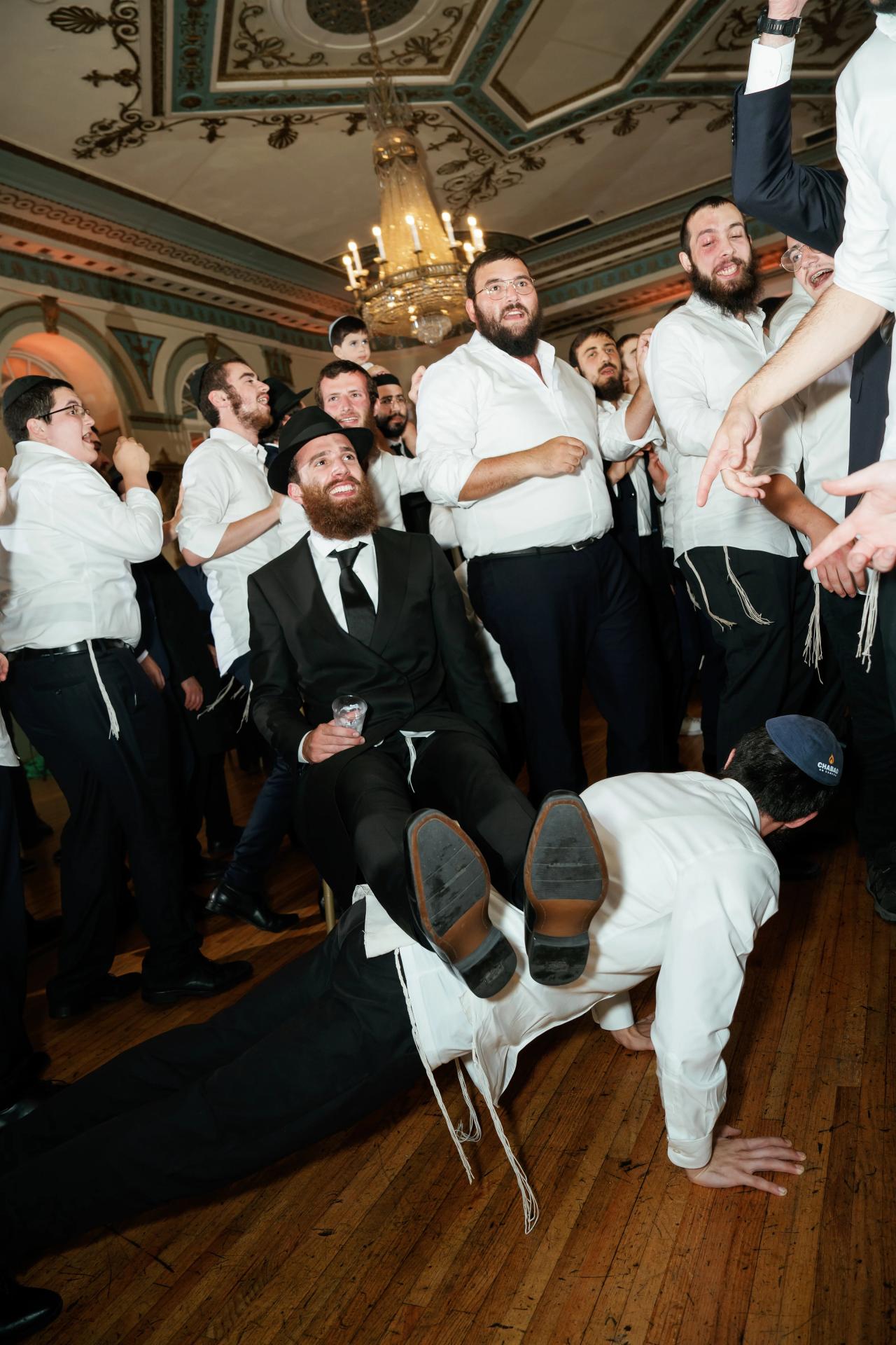 A guest performs an acrobatic floor dance in front of the smiling groom as other men cheer around them. From this Oholei Torah Ballroom wedding. Wedding photography.