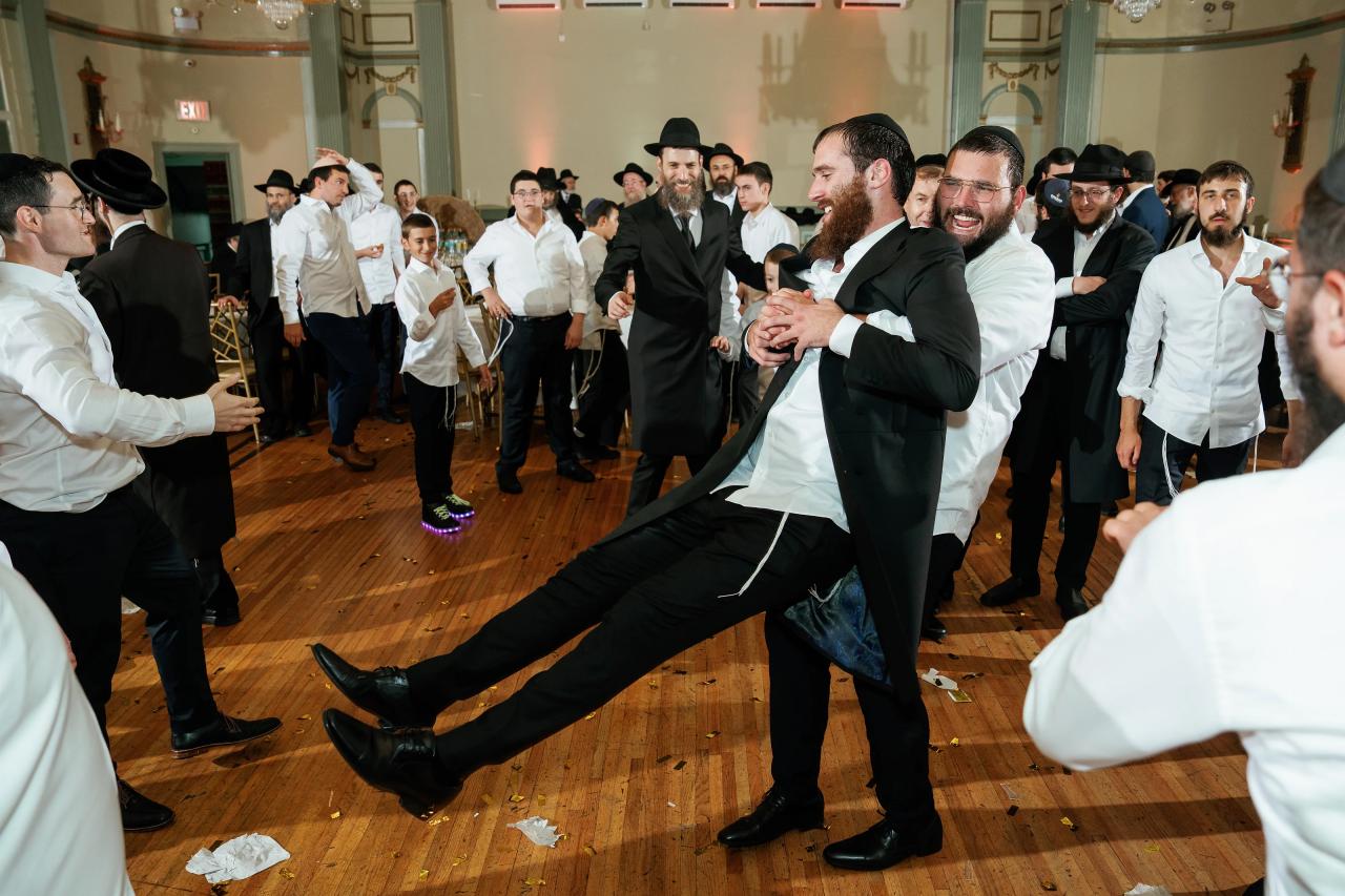 A laughing guest lifts the groom off the floor as men celebrate around them. From this Oholei Torah Ballroom wedding. Wedding photography.