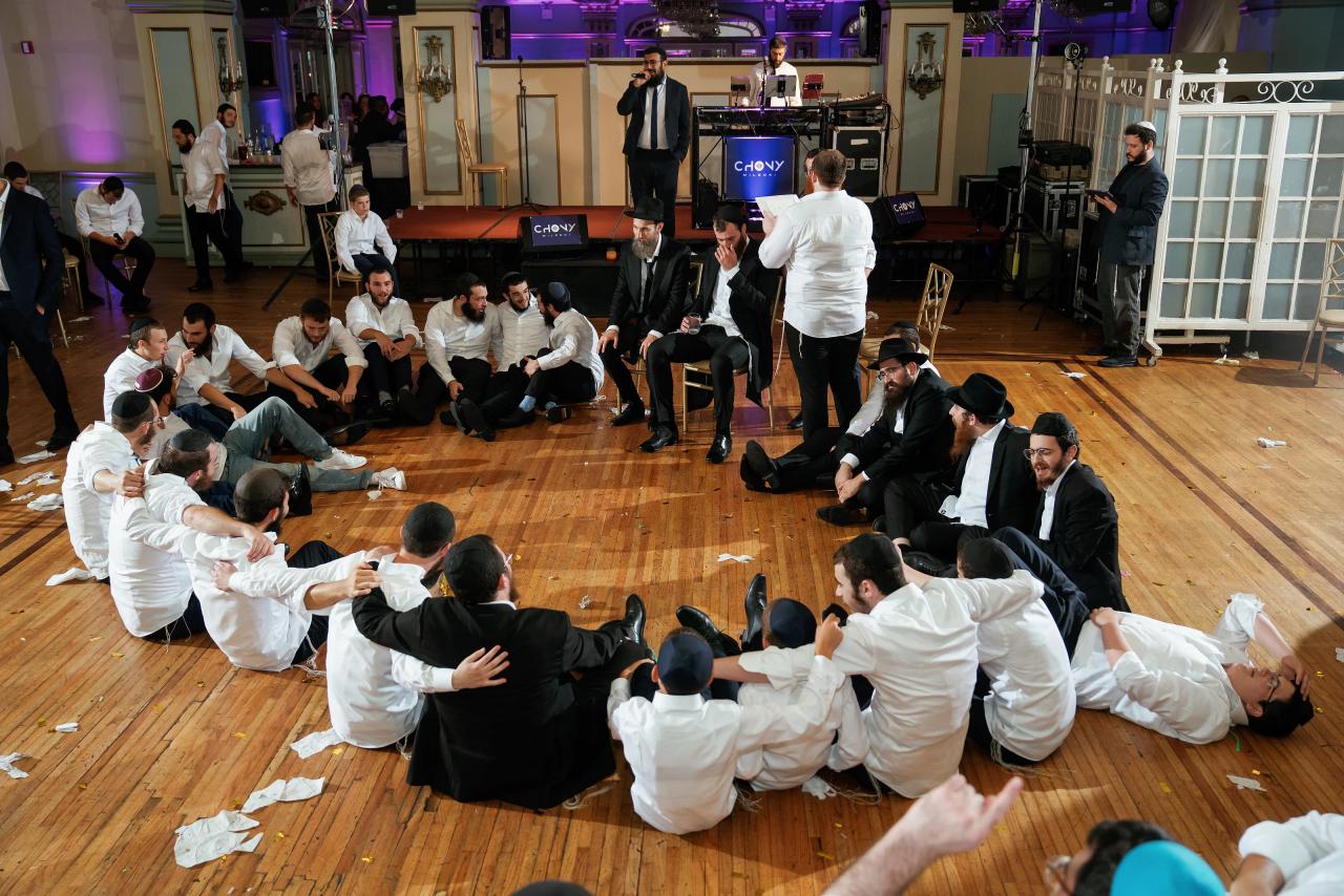 Men sit shoulder to shoulder in a large circle around the groom while musicians perform on the stage behind them. From this Oholei Torah Ballroom wedding. Wedding photography.
