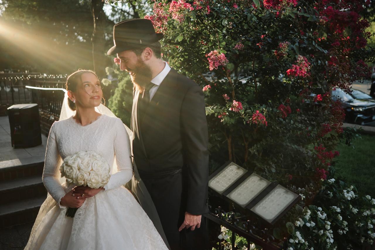 The bride looks up at the groom beside pink flowering branches as warm sunlight streams across them. From this Oholei Torah Ballroom wedding. Wedding photography.