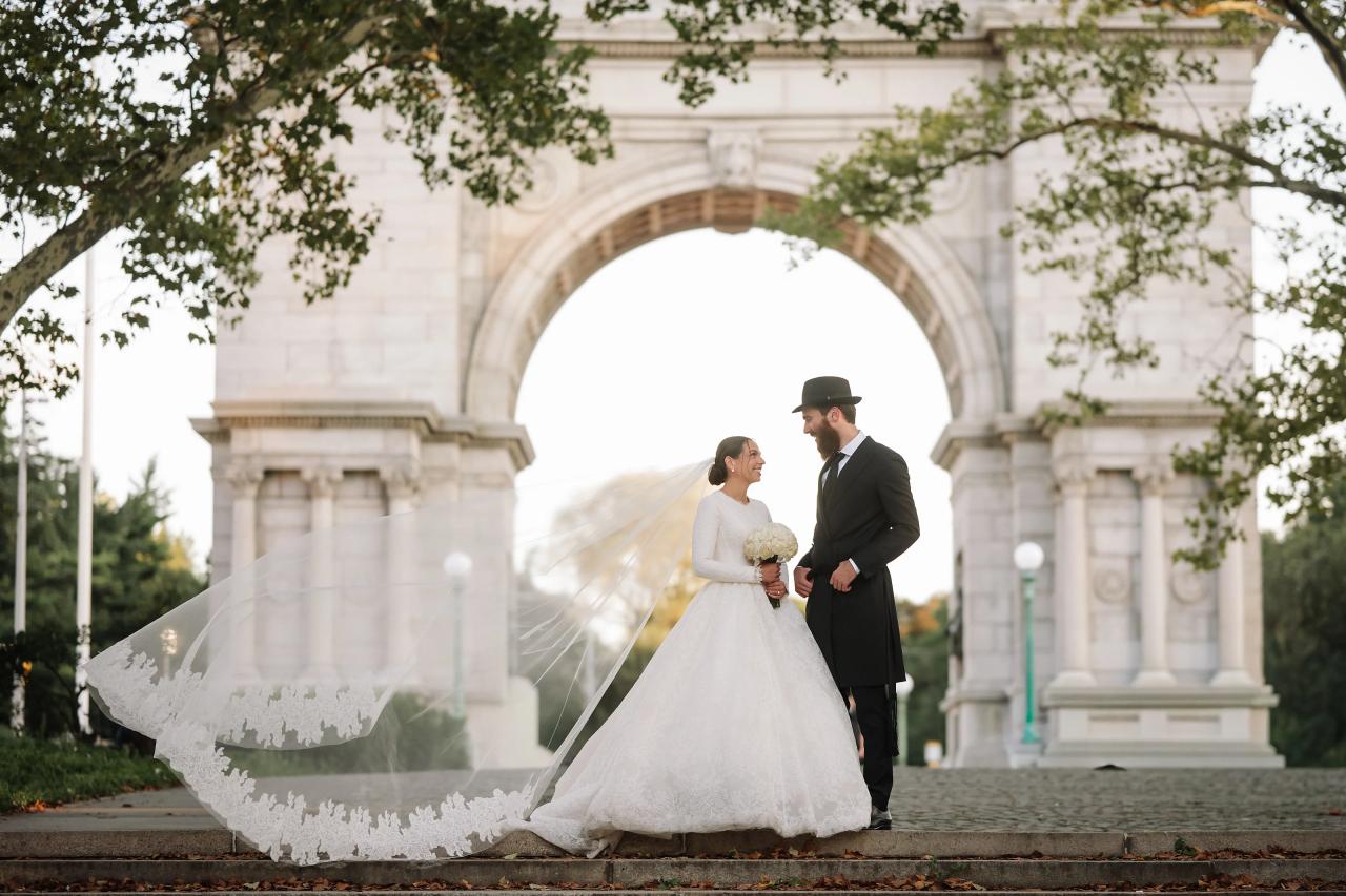 The bride and groom look toward each other in front of a monumental stone arch while her lace-edged veil sweeps to the side. From this Oholei Torah Ballroom wedding. Wedding photography.