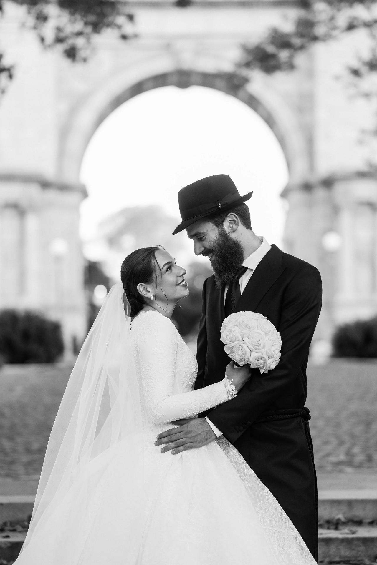 Black-and-white portrait of the bride smiling up at the groom as he holds her close, with a softly focused stone arch behind them. From this Oholei Torah Ballroom wedding. Wedding photography.