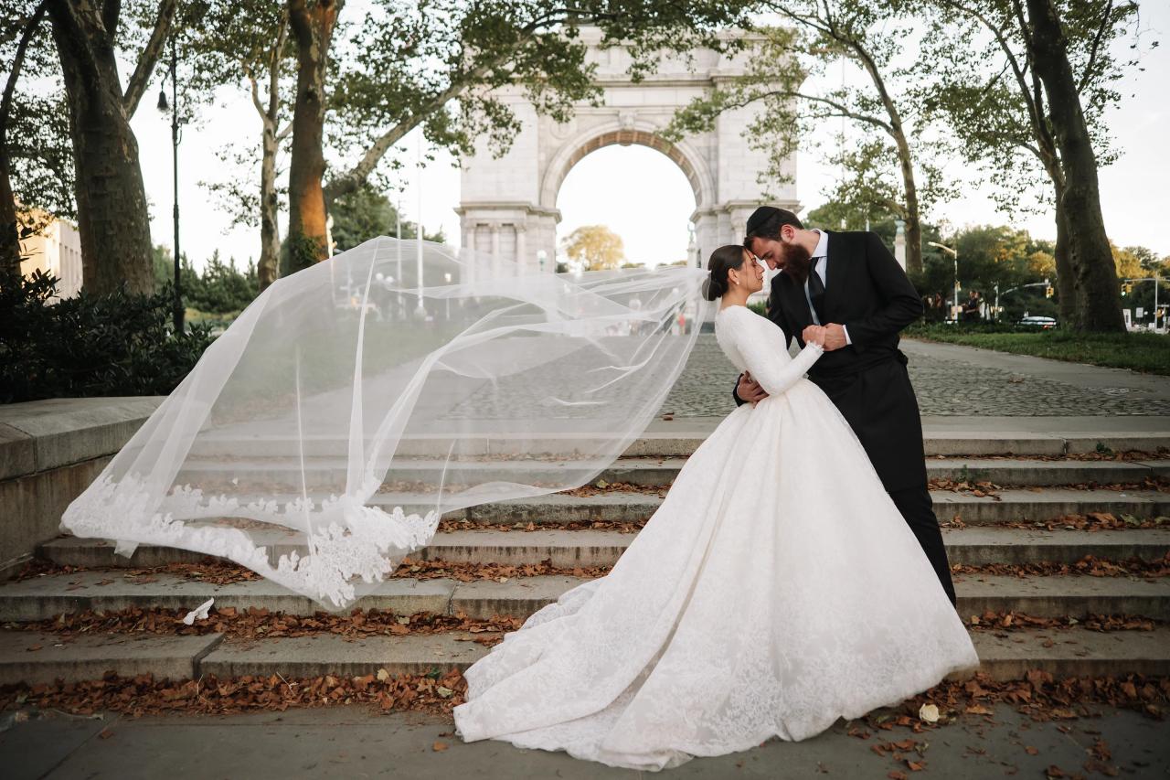 The bride and groom touch foreheads on outdoor stone steps as her long lace-edged veil billows beside them beneath the trees. From this Oholei Torah Ballroom wedding. Wedding photography.