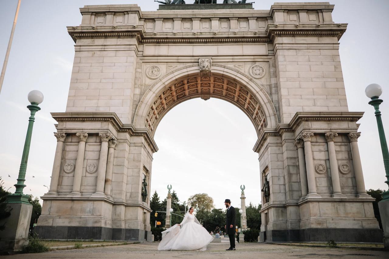The bride turns toward the groom beneath a monumental stone arch, with her white skirt moving across the paving. From this Oholei Torah Ballroom wedding. Wedding photography.