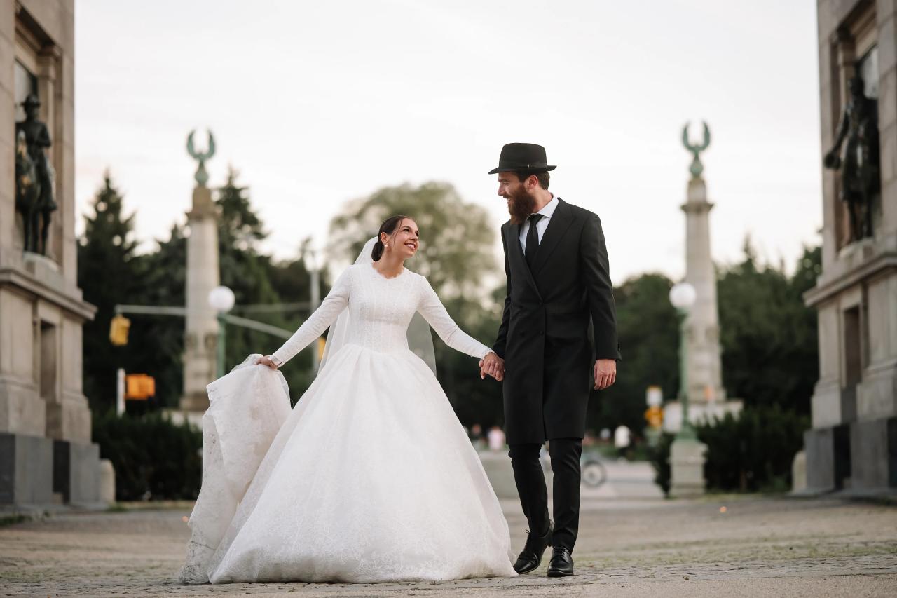The bride and groom smile at each other as they walk hand in hand through a stone archway. From this Oholei Torah Ballroom wedding. Wedding photography.