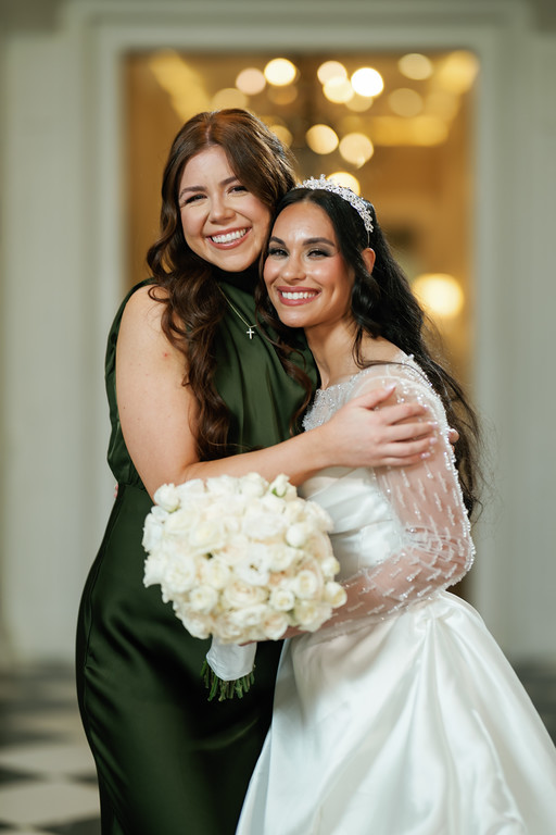 Two women in formal attire embrace and smile, with one holding a bouquet, indoors at a wedding or mitzvah celebration.