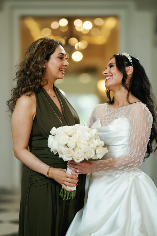 Bride in a white wedding dress holding a bouquet of white roses, smiling at a bridesmaid in a dark green dress as they stand face-to-face.