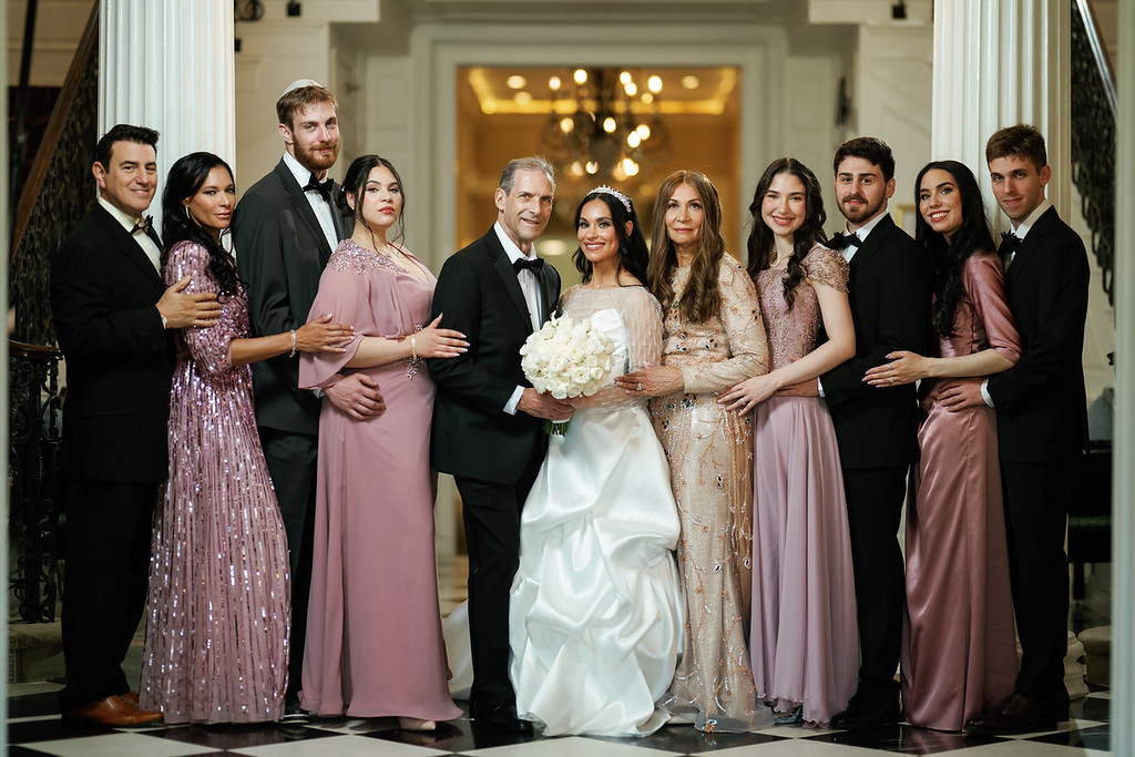 Bride standing center in a white wedding dress holding a bouquet, surrounded by family and friends in formal attire in front of a grand doorway.