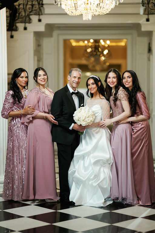Framed formal group portrait of a wedding couple standing center with family members surrounding them, smiling indoors under a chandelier.