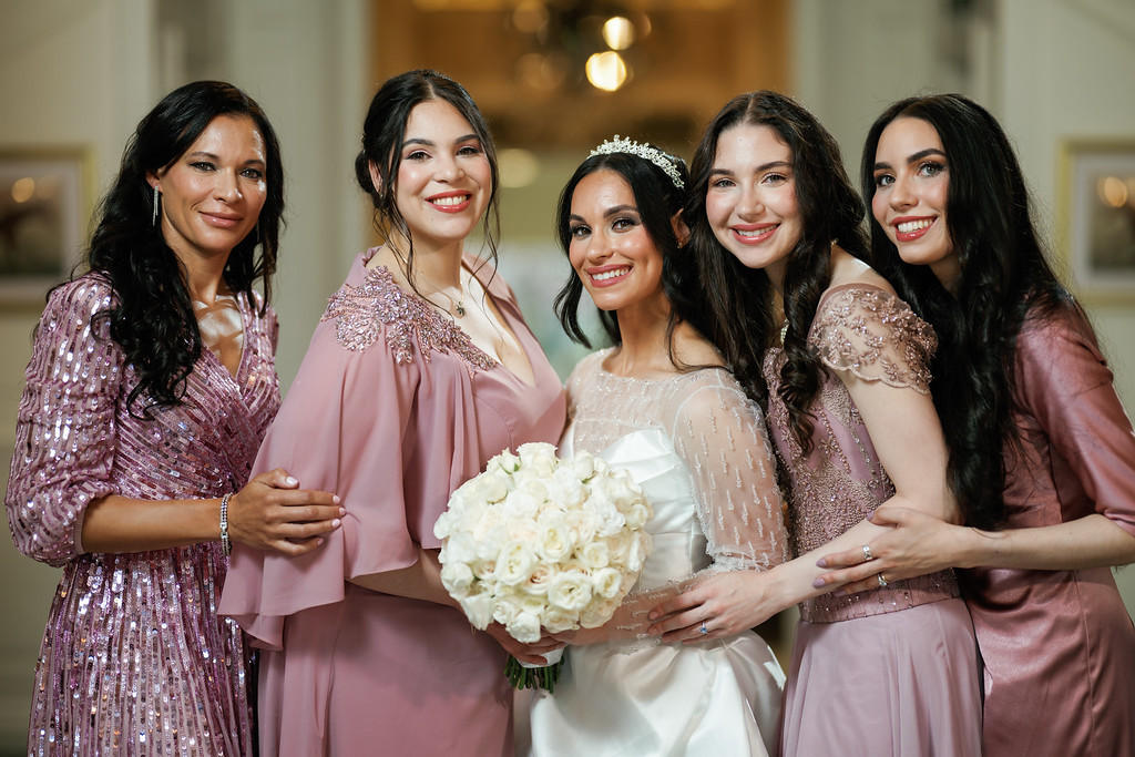 Bride holding a white bouquet while five friends in coordinated rose-pink dresses pose together, smiling in a softly lit indoor setting.
