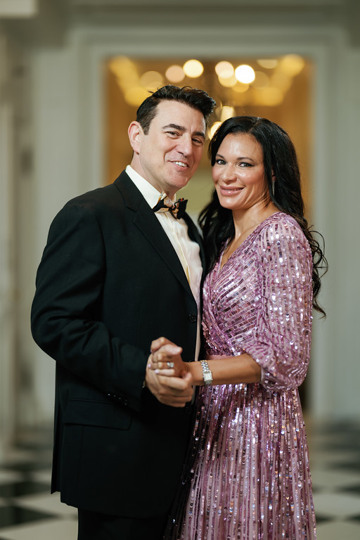 Smiling wedding couple posing together indoors, the groom in a black tuxedo with bow tie and the bride in a pink sequined dress, with warm lights blurred behind them.