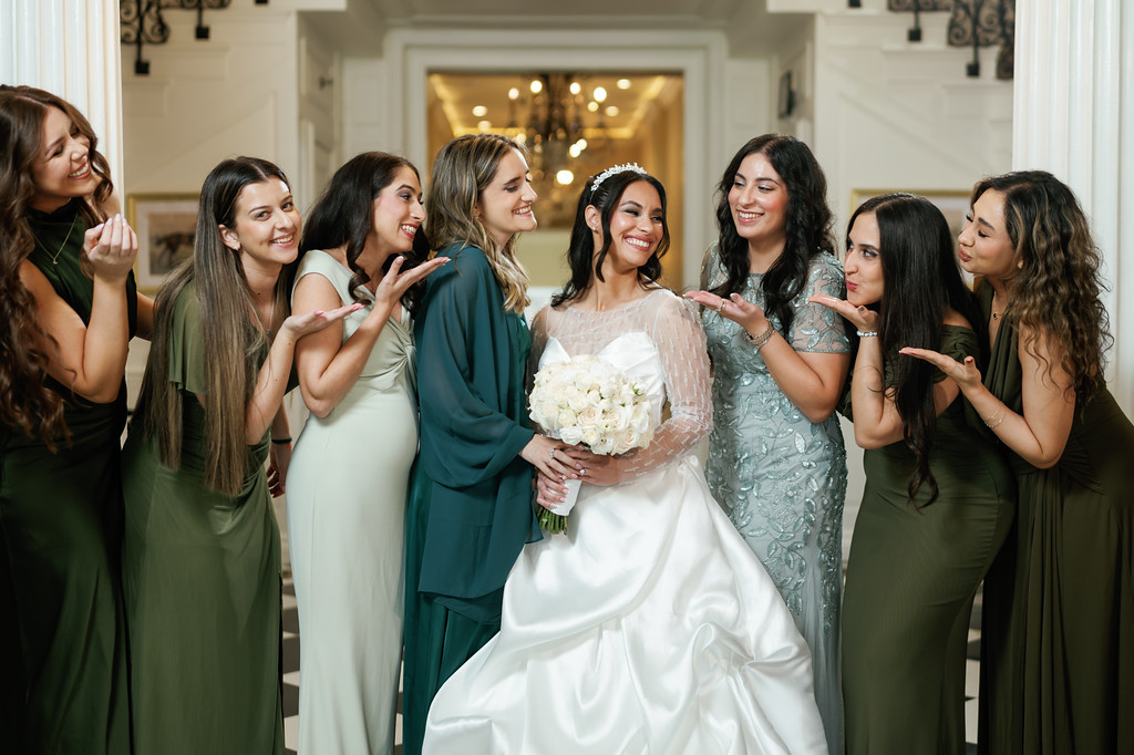 Bride smiling while her bridesmaids and friends in green dresses cheer around her with playful hand gestures during the celebration.