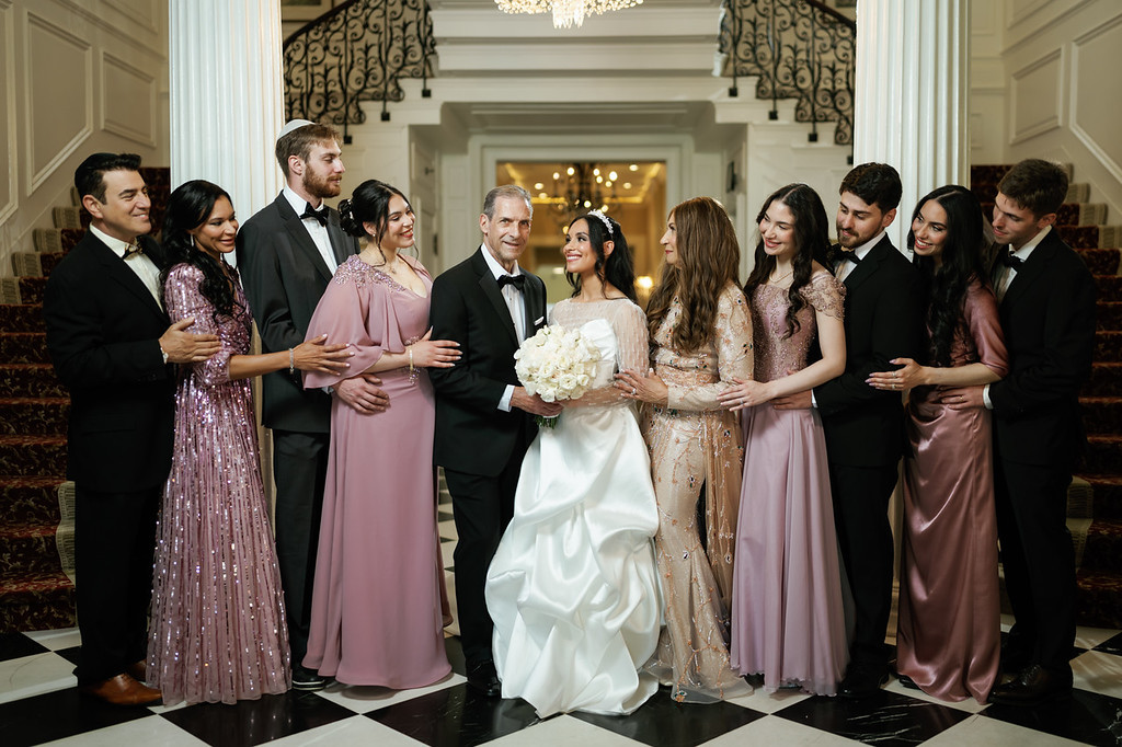 Bride in a white gown and groom in a black tuxedo stand together at the venue entrance as guests in formal attire smile and reach in for a group moment.
