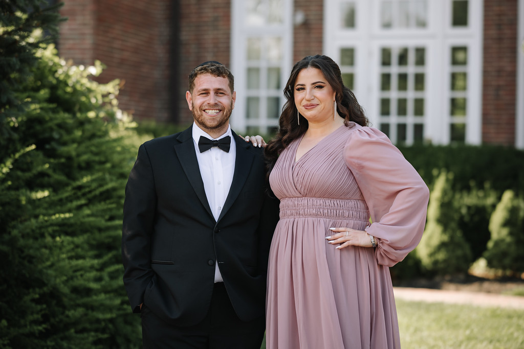 Smiling bride in a mauve dress and groom in a black tuxedo standing together outdoors in front of a brick building and manicured greenery.