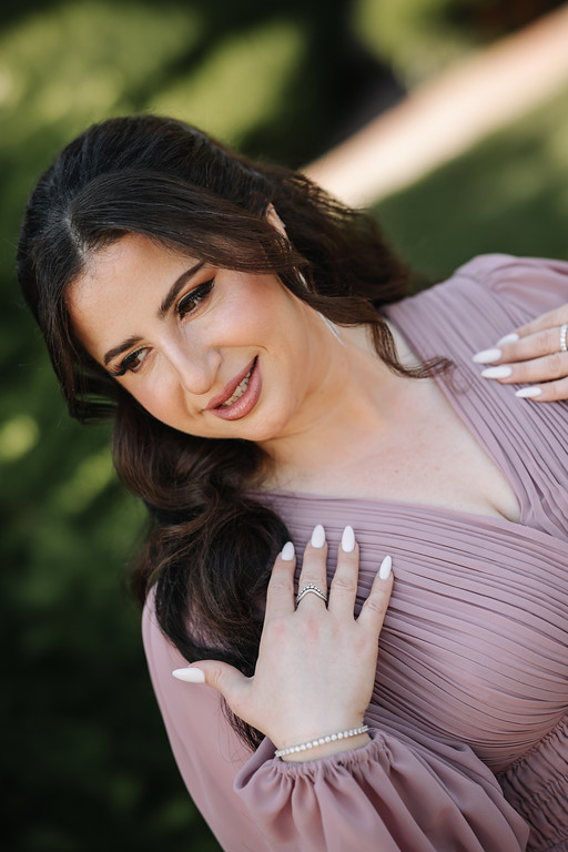 Bride in a soft pink gown with dark hair styled in loose waves, smiling as she rests her hands on her dress while posing outdoors in front of greenery.