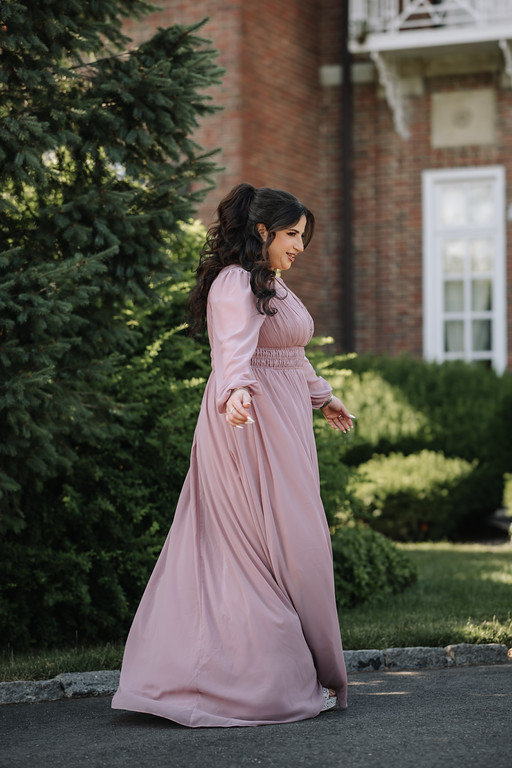 Woman in a pink wedding dress standing in a garden walkway, looking to the side with a soft expression as greenery and a brick building sit in the background.