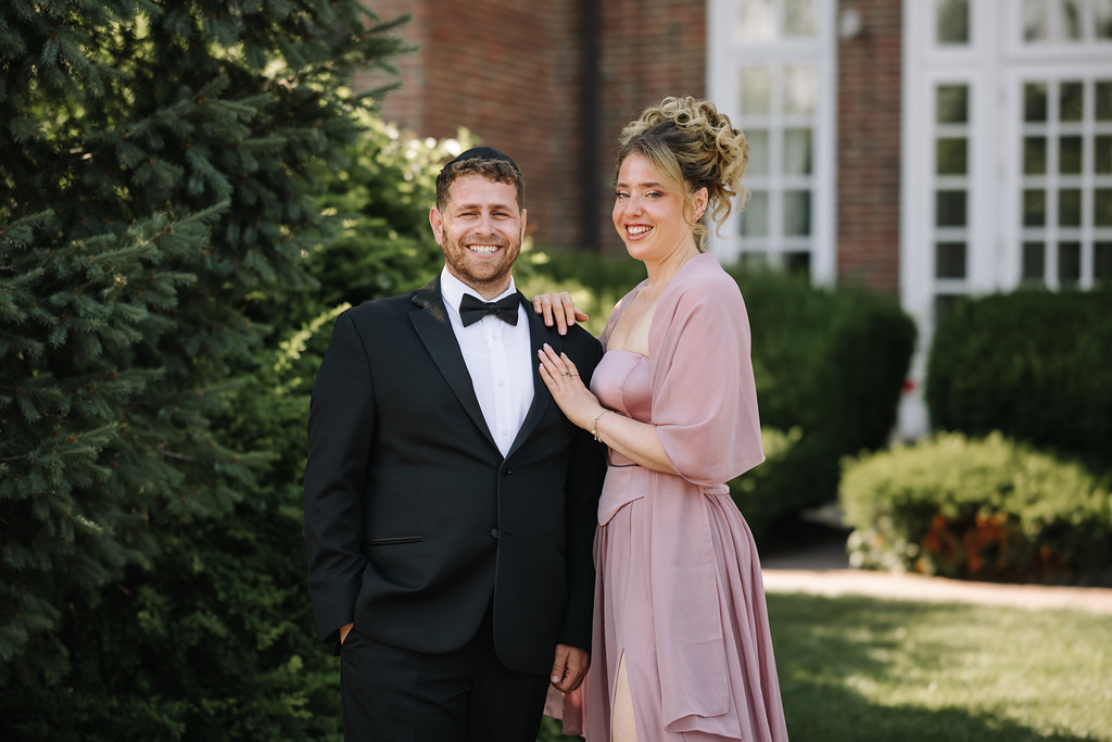 Outdoor portrait of a smiling groom in a black tuxedo and his partner in a blush dress, standing together near greenery and a brick building.