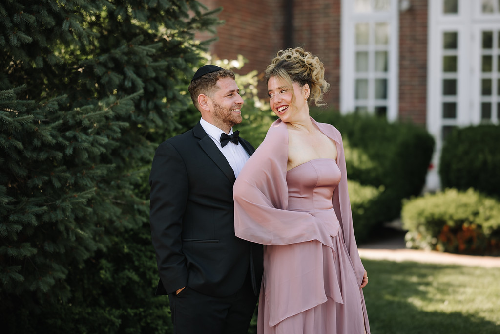Wedding couple outdoors in formal attire, standing close and smiling as they face each other in soft evening light.