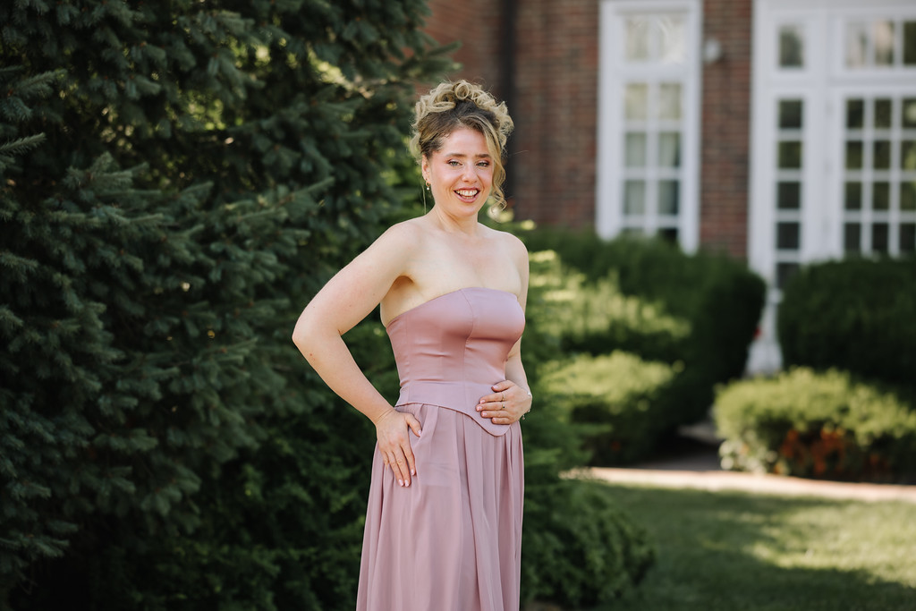 Young woman in a strapless blush dress stands outdoors in a courtyard with brick buildings and greenery behind her, smiling and looking at the camera.