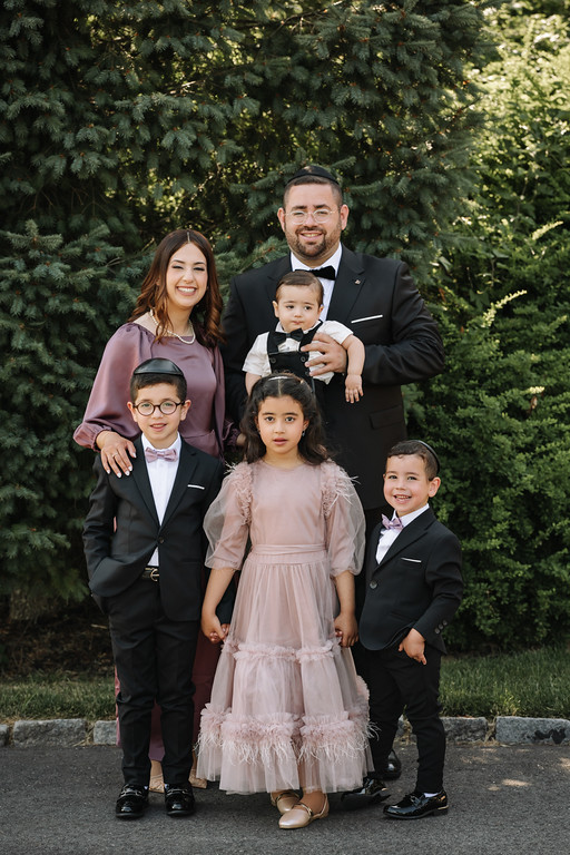Frum wedding/mitzvah-style family portrait outdoors: a father in a black tux holding a toddler, with three children in formal attire smiling in front of lush green trees.