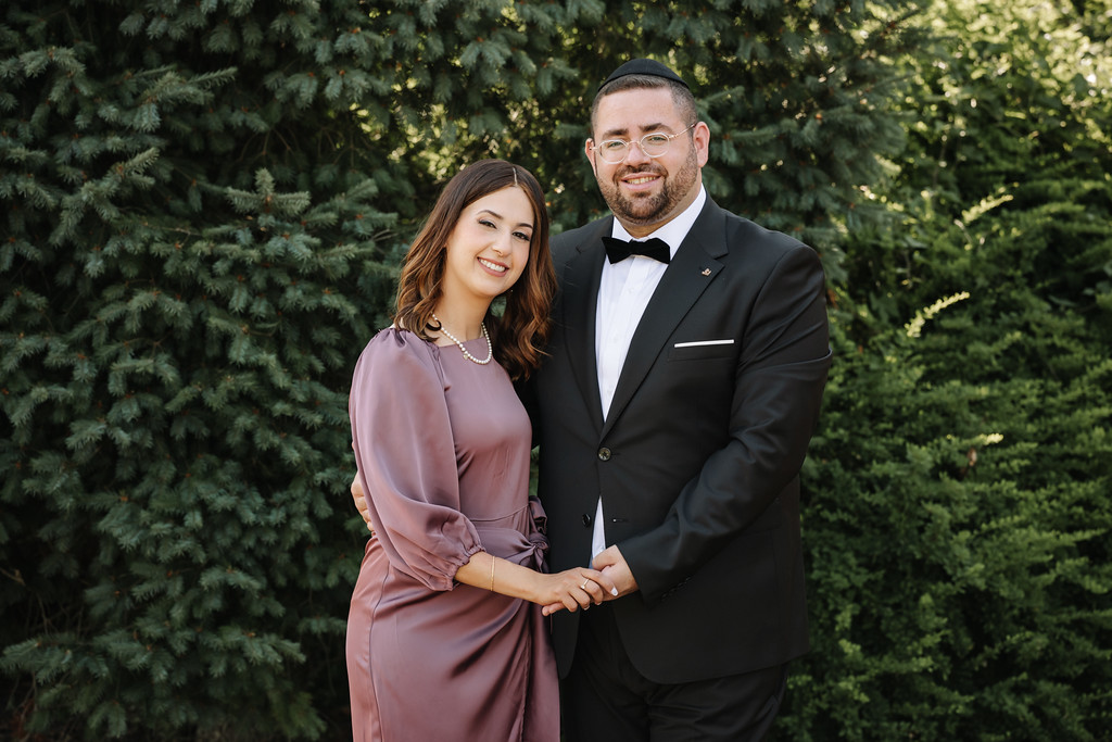 A smiling bride and groom stand together outdoors in front of evergreen trees, holding hands; the groom wears a black kippah and bow tie, and the bride wears a mauve dress.