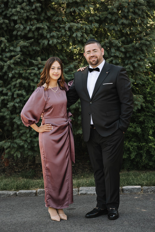 A man in a black tuxedo and bow tie and a woman in a satin mauve dress stand smiling side by side outdoors in front of evergreen trees.