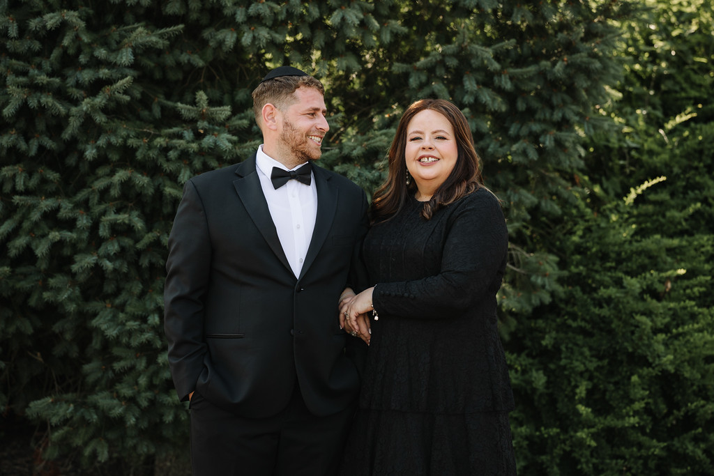 A smiling bride and groom holding hands outdoors in front of evergreen trees, dressed for their wedding ceremony.