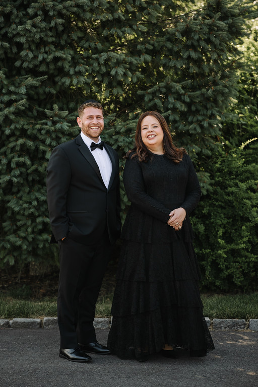 Frum wedding portrait of a groom in a black tuxedo and a bride in a dark dress, standing and smiling outdoors in front of trees.