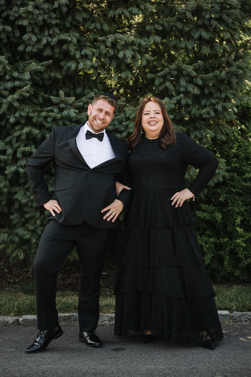 Framed outdoor portrait of a smiling groom in a black tuxedo and bow tie and a smiling bride in a black dress, standing back-to-back against green trees.