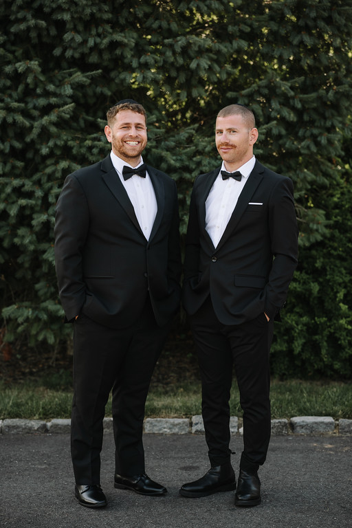 Two grooms in matching black tuxedos with bow ties stand outdoors side-by-side, smiling in front of green trees.