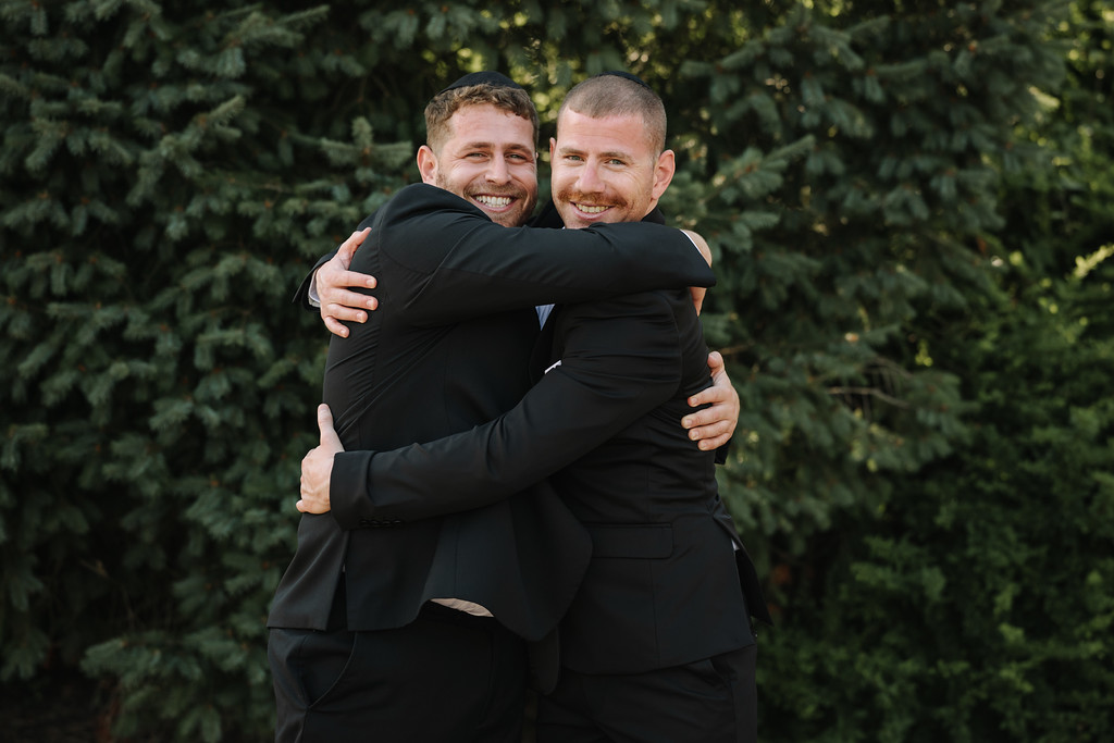 Two men in dark suits embrace warmly outdoors, smiling and laughing with arms wrapped around each other.