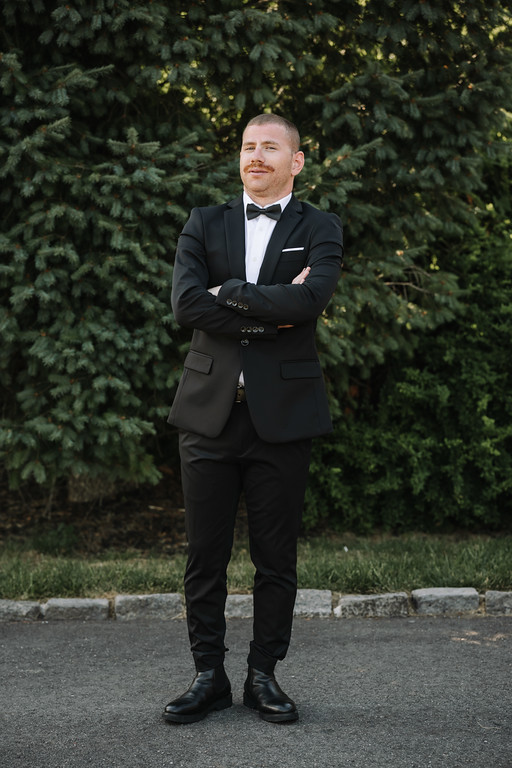 Man in a black tuxedo with a bow tie stands with arms crossed outdoors in front of evergreen trees.