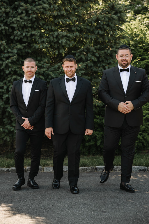 Three men in black tuxedos with bow ties walking toward the camera outdoors in front of green trees, one smiling in the center.