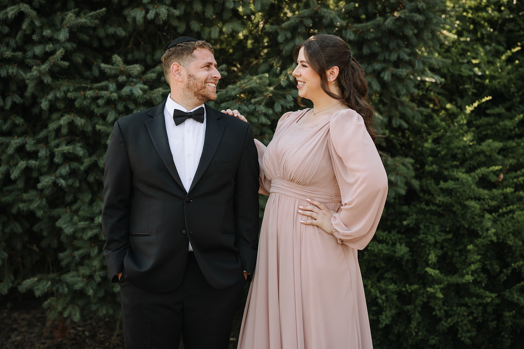 A groom in a black tuxedo and a bride in a light pink gown stand outdoors, smiling and looking at each other against a backdrop of evergreen trees.