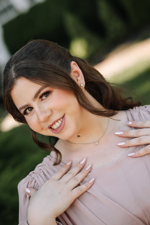 Smiling bride in a soft embrace outdoors, her hands resting on her partner’s shoulder with a garden background in soft focus.