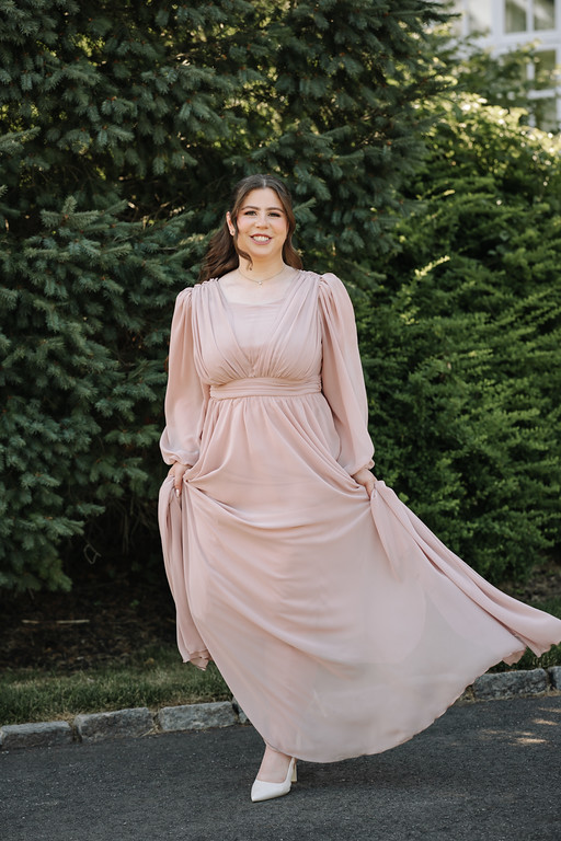 Portrait of a smiling bride in a blush-pink dress standing outdoors in front of evergreen trees, her dress flowing in the breeze.