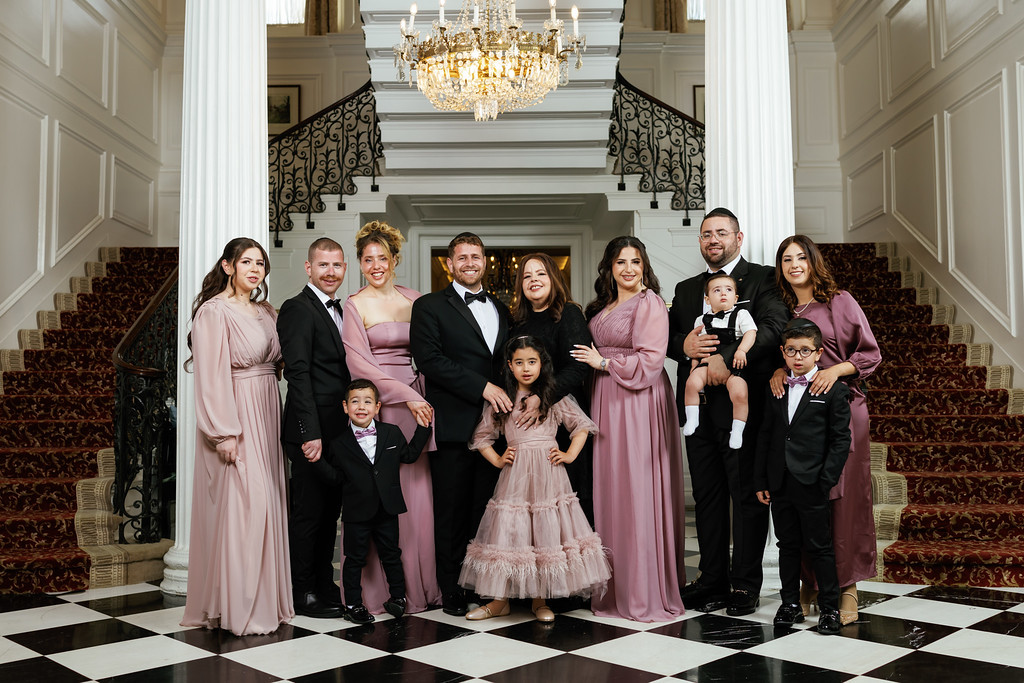 Family portrait in a formal ballroom with a chandelier overhead, dressed in coordinated mauve and black outfits, standing together on a checkered floor.