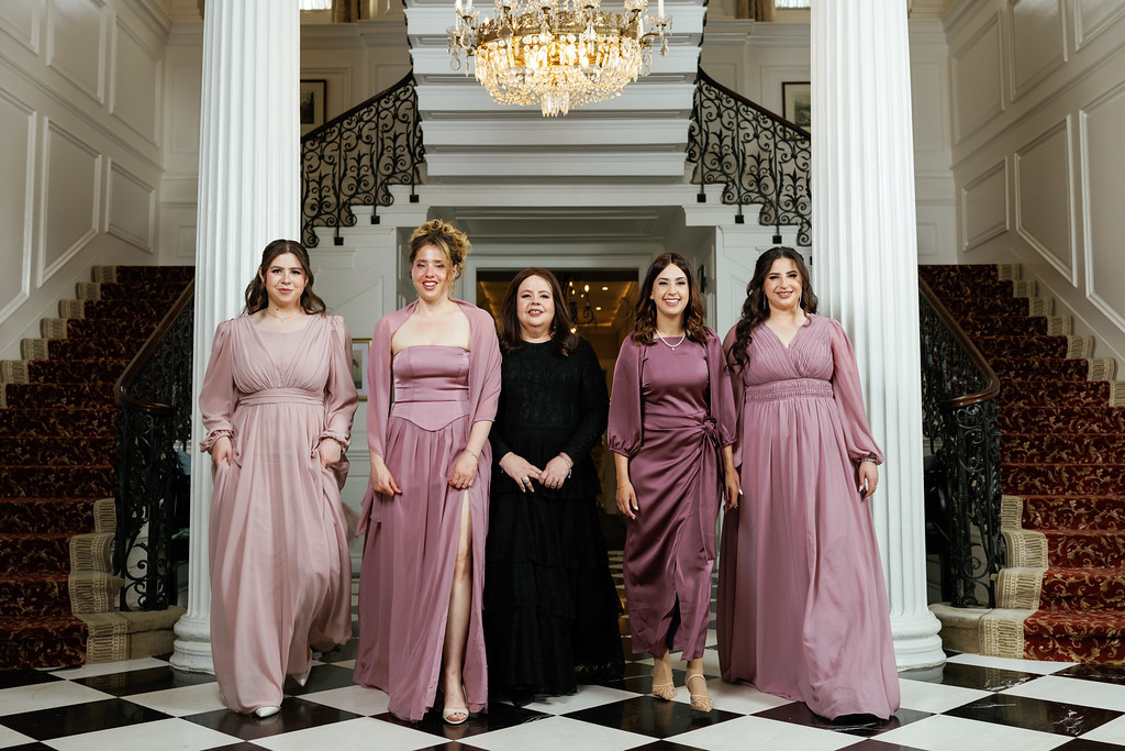 Five women wearing coordinated pink dresses pose together for a formal group photo in a grand hall with white columns and a chandelier overhead.