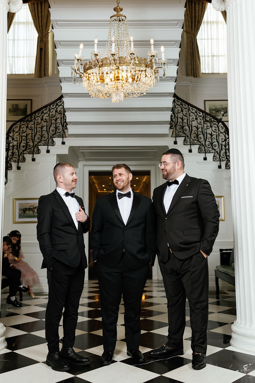 Three men in black tuxedos with bow ties stand together indoors under a chandelier, smiling and conversing at a formal event.