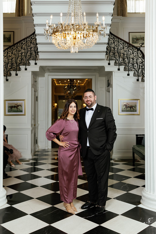 Frum newlywed couple posing side by side in a grand ballroom entrance with a chandelier overhead, black-and-white checkered floor, and matching formal outfits.