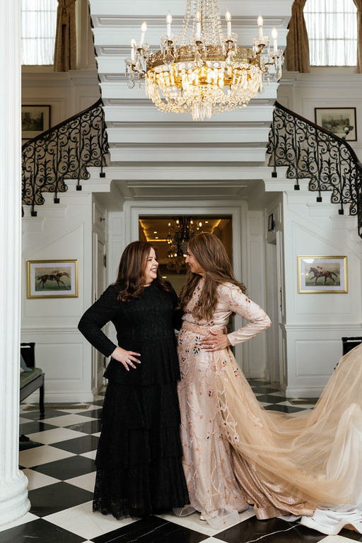 Two women in formal dresses stand face-to-face inside a grand white foyer with a chandelier above, smiling and posing.