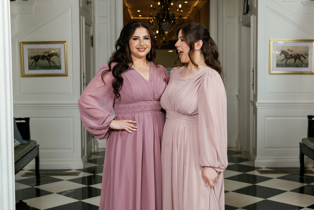 Two women in pink dresses smiling and laughing together in a well-lit hallway during a Jewish celebration.